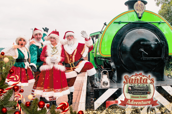 Festive group in Santa attire poses by a green steam train with Christmas decorations.